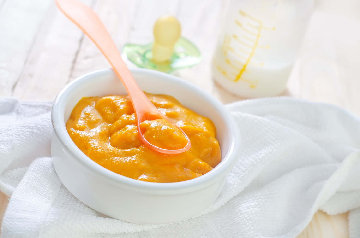Bowl of orange baby puree with spoon on a white cloth, with a baby bottle and pacifier in the background