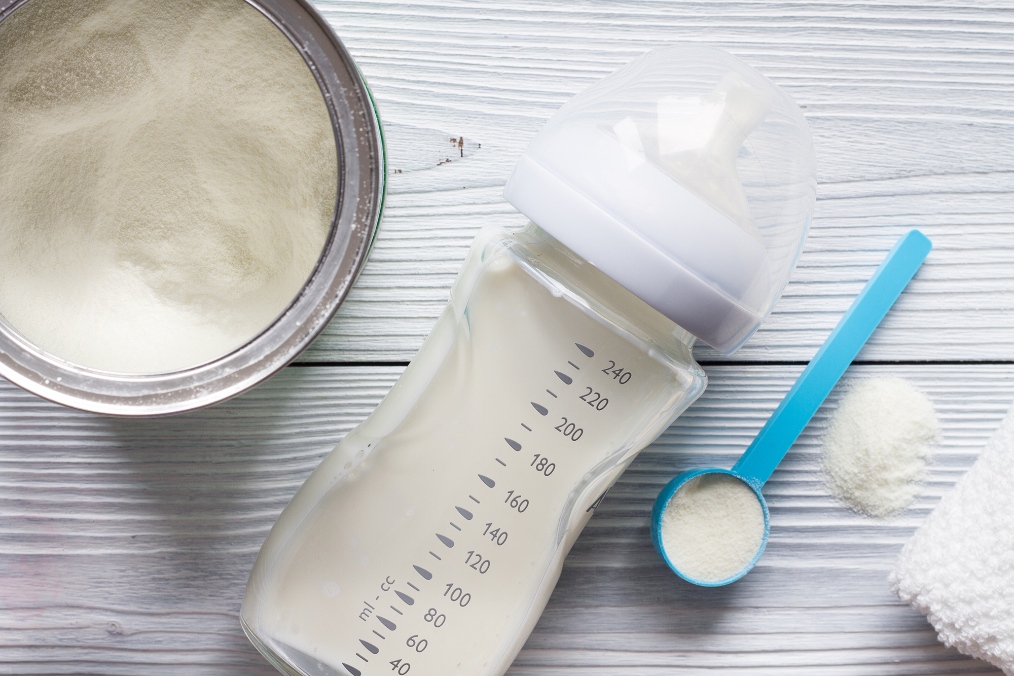 Baby bottle with milk and measurement markings next to a blue scoop with powdered formula, an open tin of powder, and a white cloth on a light wooden table
