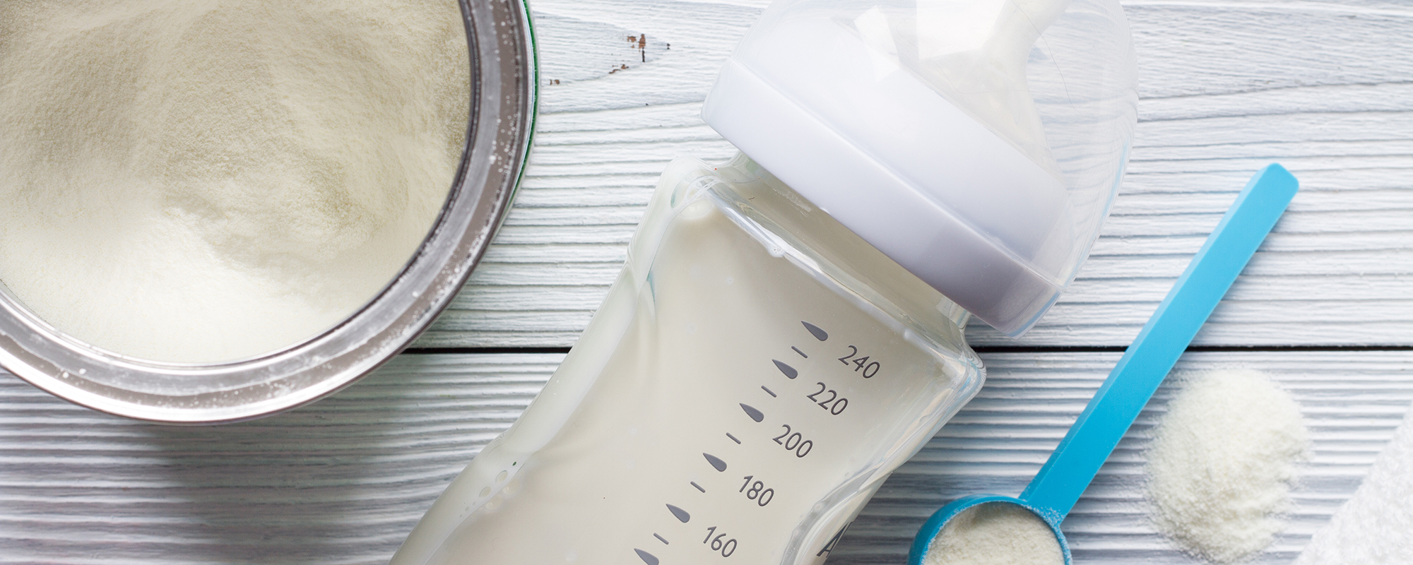 Baby bottle with milk and measurement markings next to a blue scoop with powdered formula, an open tin of powder, and a white cloth on a light wooden table